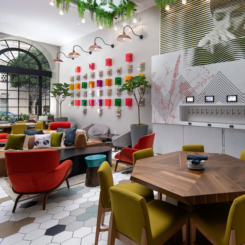 A cozy cafe with colorful lounge chairs, a round wooden table, warm lighting, and shelves of bottles against a glass wall.