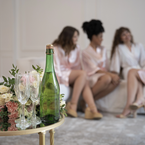 A champagne bottle with flutes and a floral arrangement on a small round table, while four women in light robes chat on a sofa in the background.