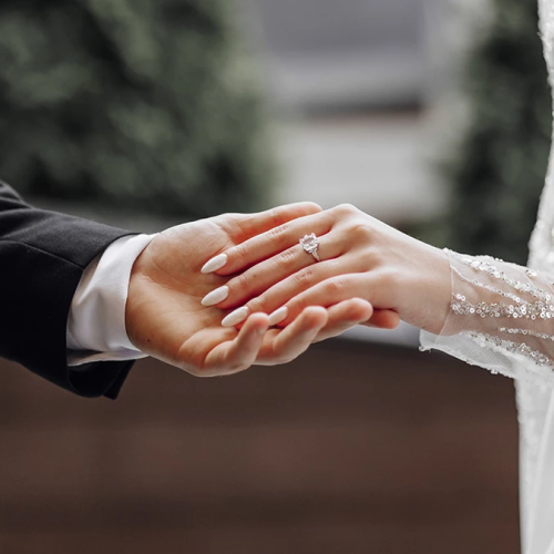 A couple holds hands, showing rings, wedding attire, and a tender moment between a bride and groom.