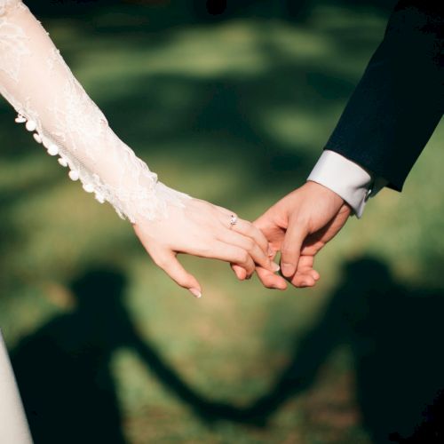 A couple holding hands, wedding attire, outdoors, symbolizing unity and vows, with soft shadows in the background.