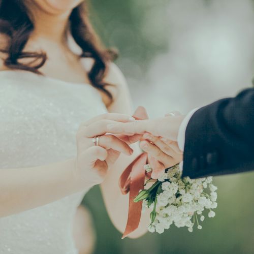 A bride and groom exchange rings during a wedding ceremony, the groom placing a ring on the bride&rsquo;s finger while she holds a small bouquet.