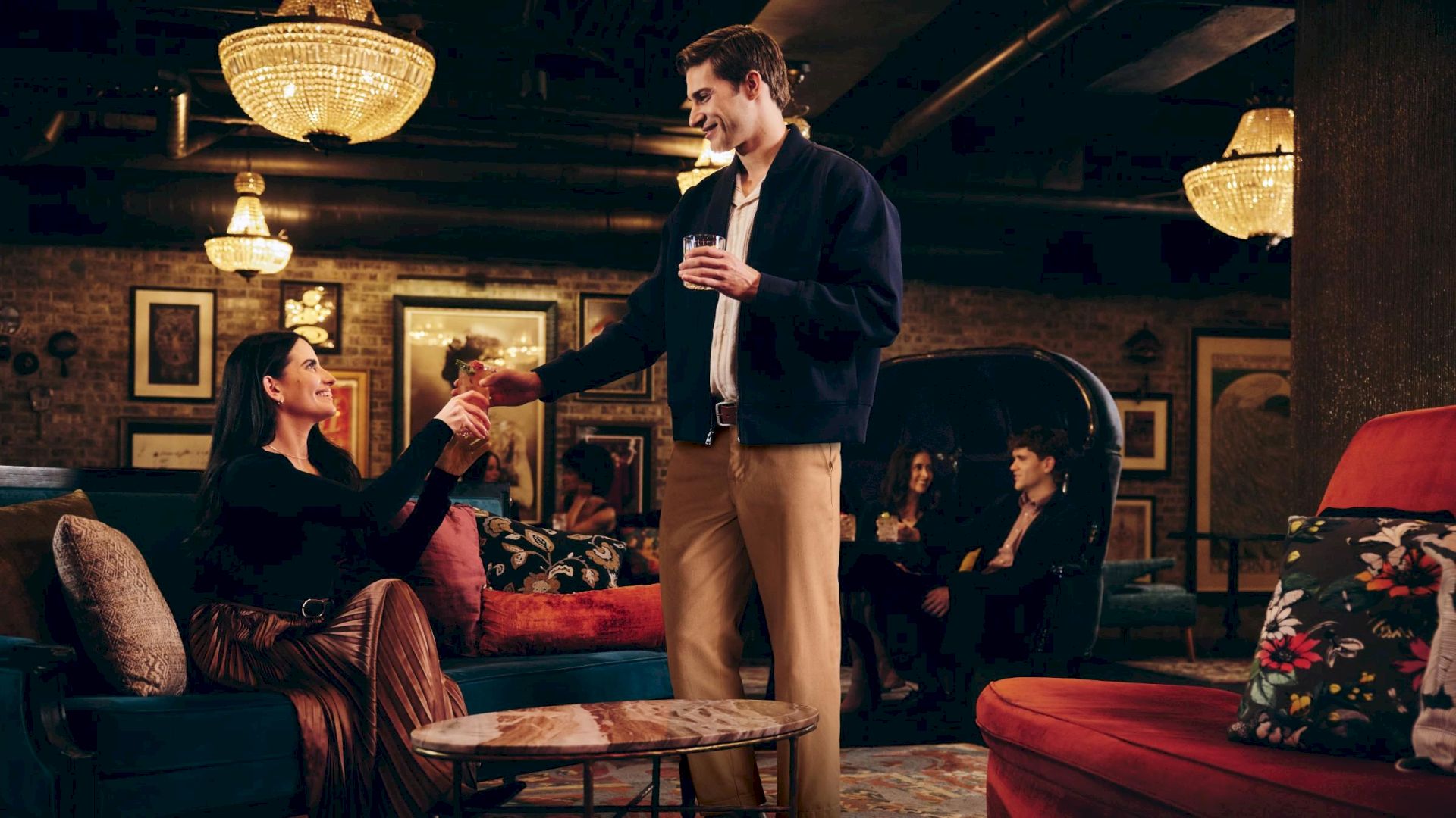 A man in a navy jacket offers a drink to a seated woman in a cozy lounge; warm lamps, other guests, and casual chatter in the background.