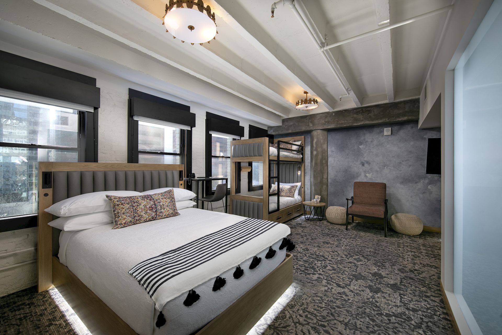 A modern hotel bedroom with a large bed, striped linens, cushioned headboard, carpeted floor, and a seating area with a chair and stone wall accents.