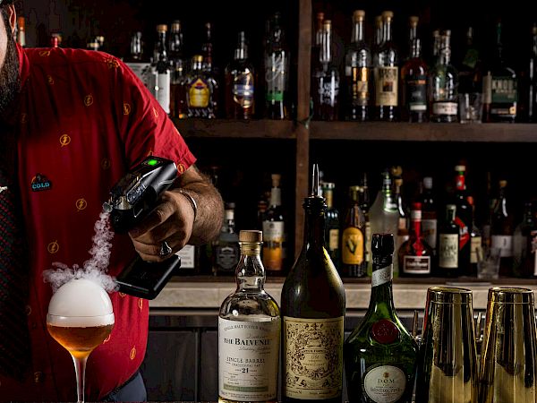 A bartender with a red shirt pours ember foam into a glass, crafting a cocktail with bottles and mixers lined up behind the bar.