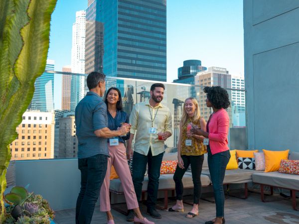 A group of five friends chat and laugh on a rooftop in a sunny city, with tall buildings in the background and colorful cushions on the seating.