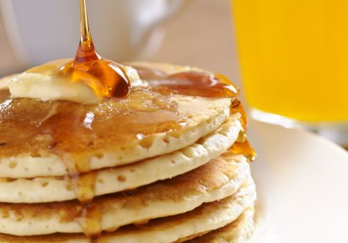 Stack of pancakes with syrup being poured, butter melting, and a glass of orange juice in the background, breakfast vibes.