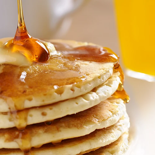 Stack of pancakes with syrup being poured, butter melting, and a glass of orange juice in the background, breakfast vibes.