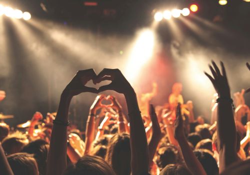 Crowd at a concert, hands raised, stage lights blaze, someone forms a heart shape with fingers&mdash;the vibe is energetic and festive. End sentence.