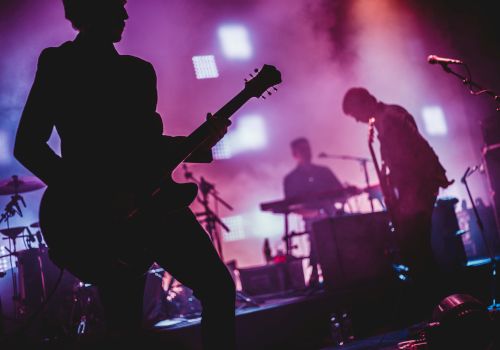 A live rock band on stage with silhouetted guitarists, bright purple lights, and a drummer in the background, creating a moody concert vibe.