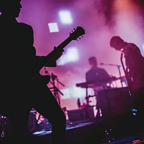 A live rock band on stage with silhouetted guitarists, bright purple lights, and a drummer in the background, creating a moody concert vibe.