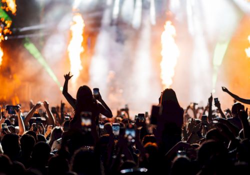 A packed concert crowd cheers with raised phones as huge flame jets shoot up onstage.