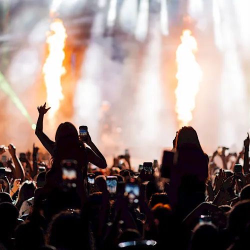 A packed concert crowd cheers with raised phones as huge flame jets shoot up onstage.