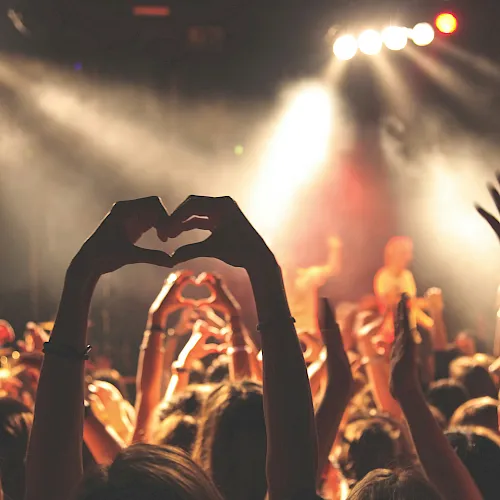A concert crowd forms heart hands toward a brightly lit stage, cheering as musicians perform.