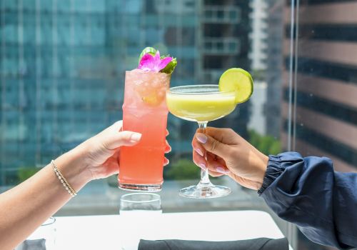 Two people clink cocktails: a pink drink with a flower garnish and a yellow drink topped with a lime slice, outdoors.