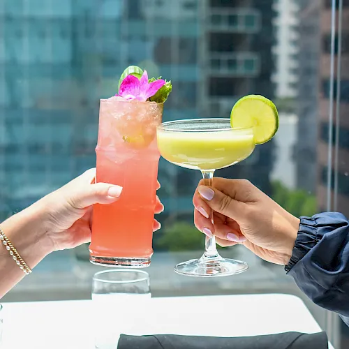 Two people clink cocktails: a pink drink with a flower garnish and a yellow drink topped with a lime slice, outdoors.