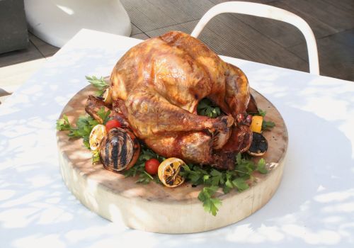 A roasted chicken on a wooden board with herbs, tomatoes, lemons, and garlic, ready to serve.